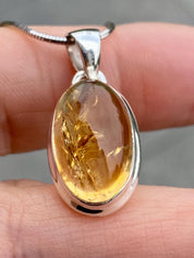 Handmade sterling silver pendant featuring an oval cabochon-cut golden yellow citrine gemstone. The bezel-set stone has natural inclusions and a smooth, polished finish. Suspended from a delicate silver chain, shown against a hand for scale.