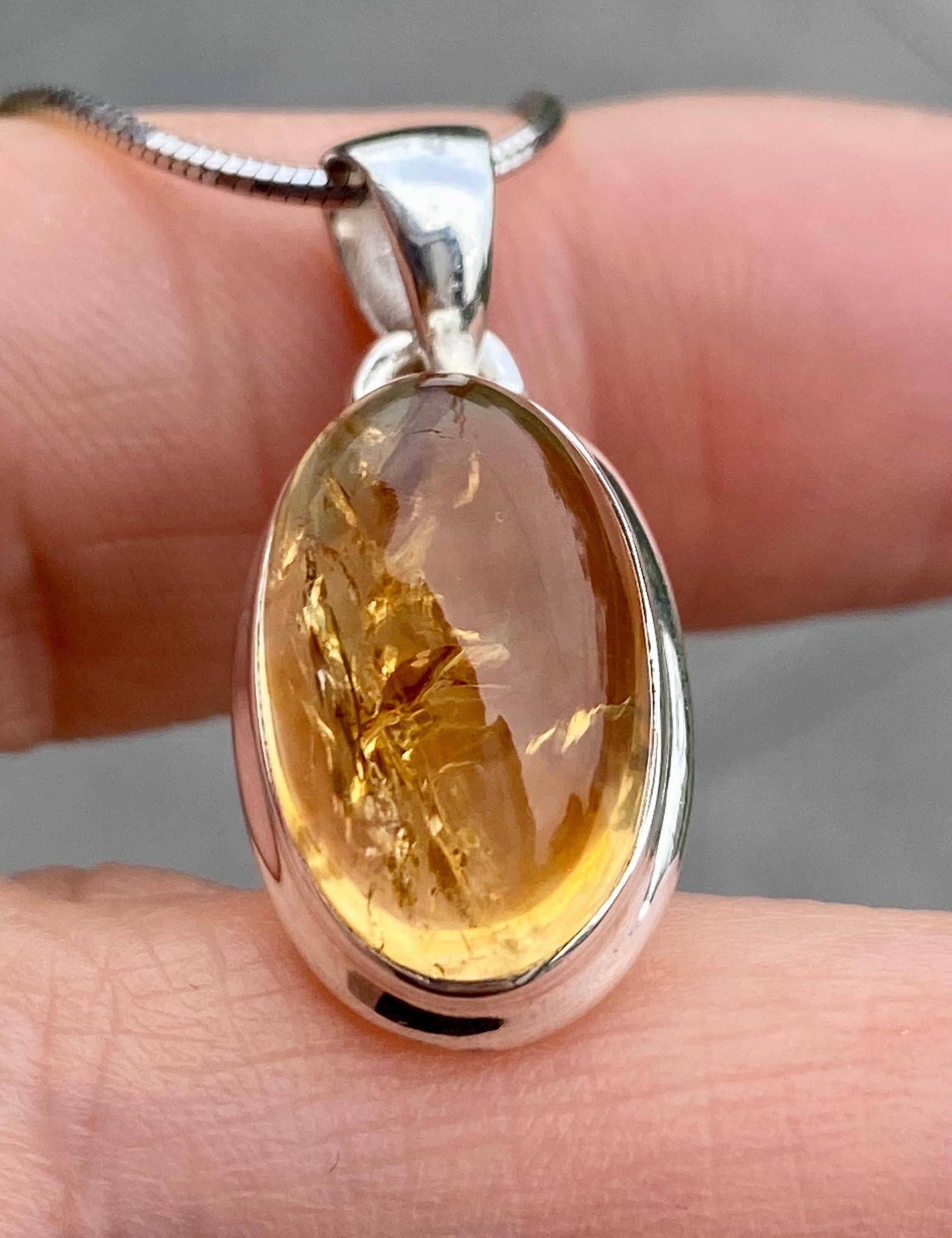 Handmade sterling silver pendant featuring an oval cabochon-cut golden yellow citrine gemstone. The bezel-set stone has natural inclusions and a smooth, polished finish. Suspended from a delicate silver chain, shown against a hand for scale.