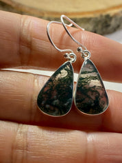 Handmade sterling silver earrings featuring teardrop-shaped Moss Agate gemstones with intricate green inclusions. The polished stones are bezel-set, hanging from elegant silver hooks. Photographed on a hand for scale and natural lighting effect.
