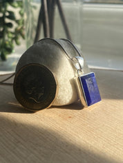 Sterling silver pendant featuring a rectangular deep blue Sapphire gemstone with a smooth finish. The pendant is displayed alongside a £1 coin for scale, resting on a natural stone with soft lighting for an elegant presentation