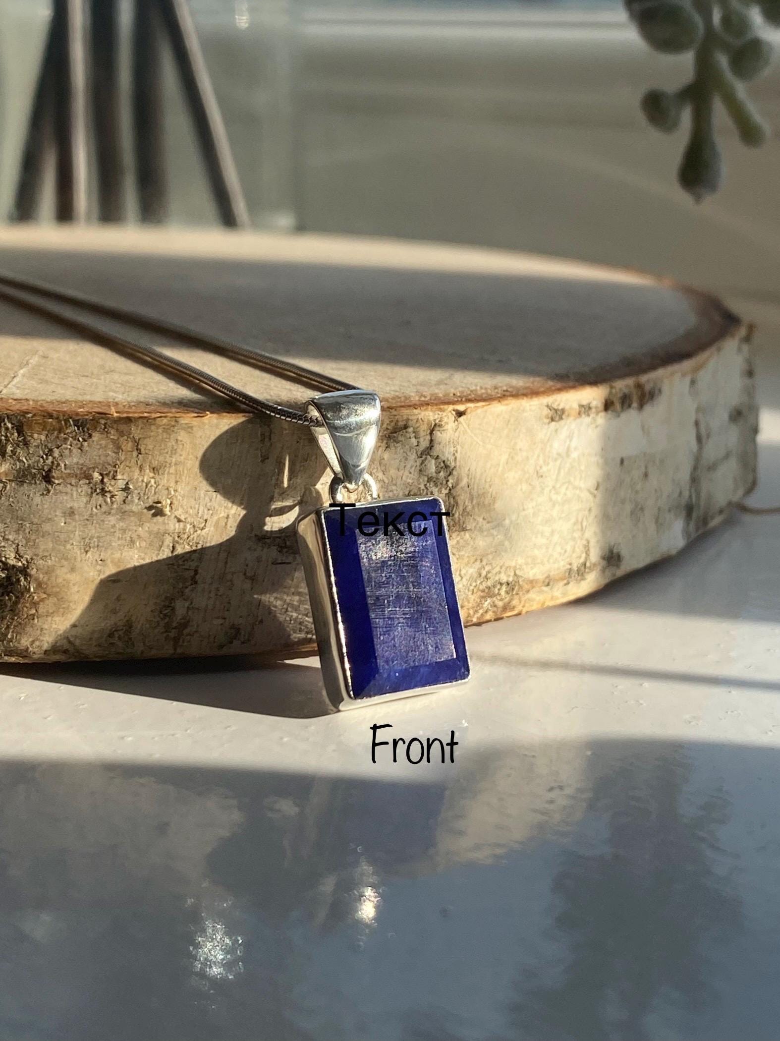 Sterling silver pendant featuring a rectangular deep blue Sapphire gemstone with a smooth, polished finish. The pendant is displayed on a wooden slice, with natural sunlight highlighting its rich colour. &#39;Front&#39; text label added for clarity.