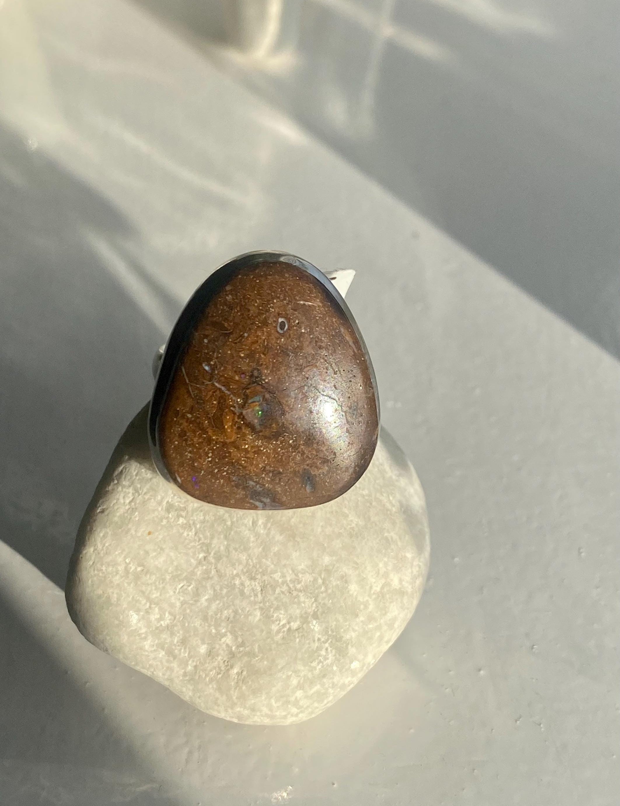 Handmade statement ring featuring a polished brown Australian boulder opal with natural inclusions, set in a sterling silver band. The stone displays a subtle play of colour under sunlight. Photographed on a smooth white stone for contrast.