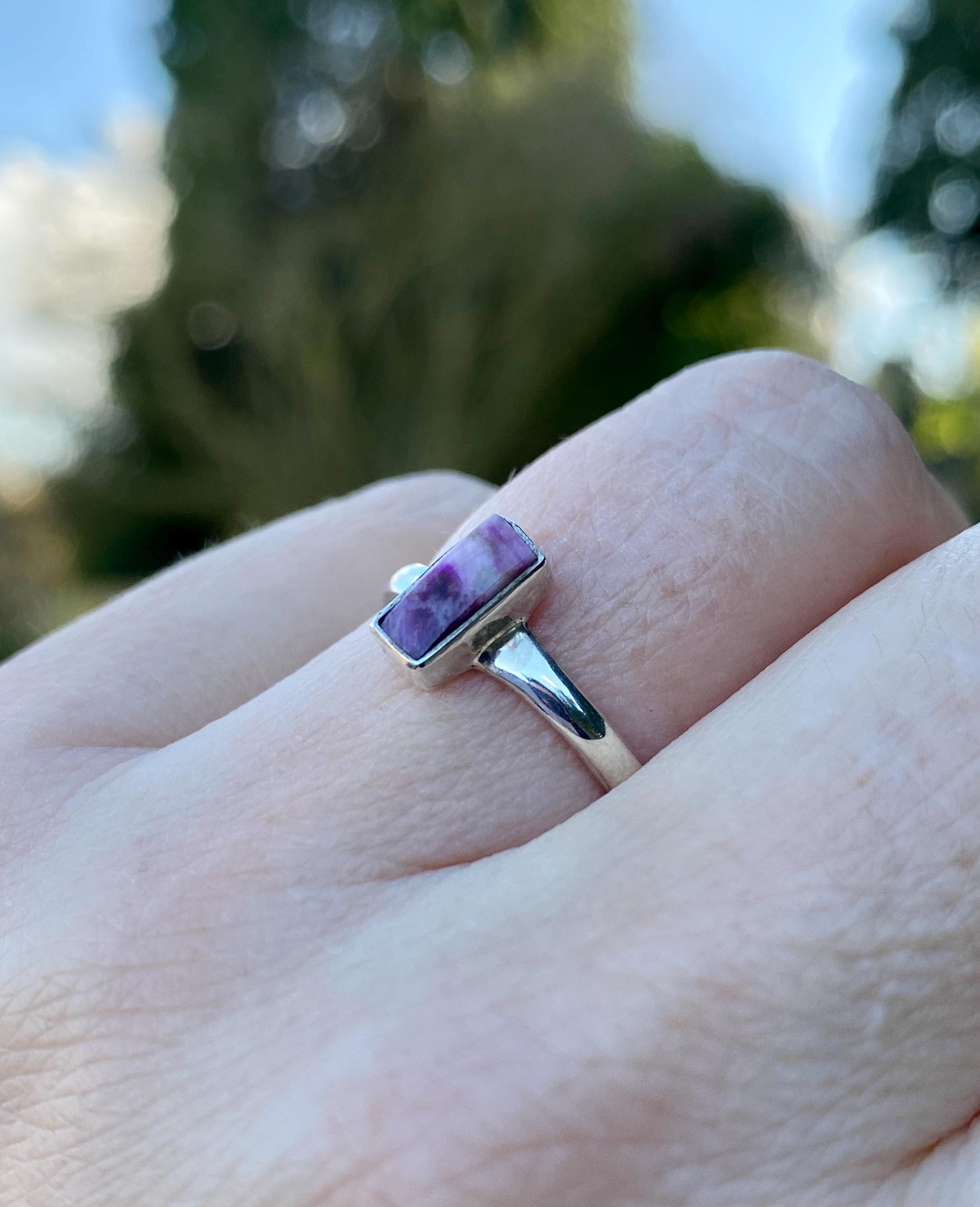 Handmade sterling silver sugilite ring featuring a rectangular purple and white gemstone with a marbled effect. The stone is bezel-set, and the band is slim and polished. Photographed outdoors on a neutral surface with a blurred natural background.