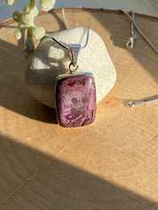 Handmade pendant featuring a rectangular, polished pink, purple, white and black Sugilite gemstone with natural patterns, bezel-set in 925 sterling silver. The pendant hangs from a delicate silver chain and is photographed in natural sunlight on a wooden surface.