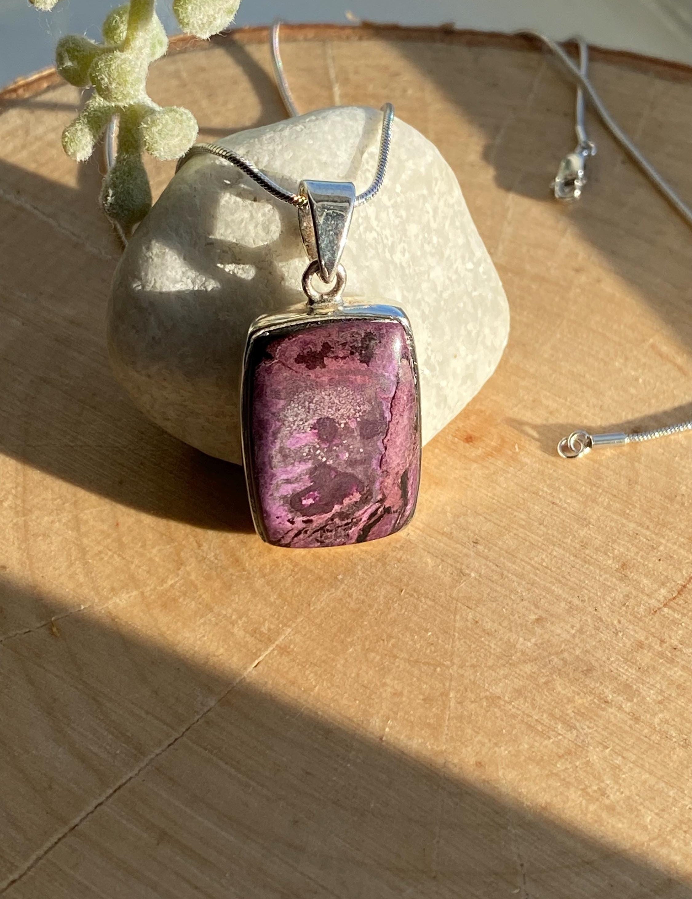 Handmade pendant featuring a rectangular, polished pink, purple, white and black Sugilite gemstone with natural patterns, bezel-set in 925 sterling silver. The pendant hangs from a delicate silver chain and is photographed in natural sunlight on a wooden surface.