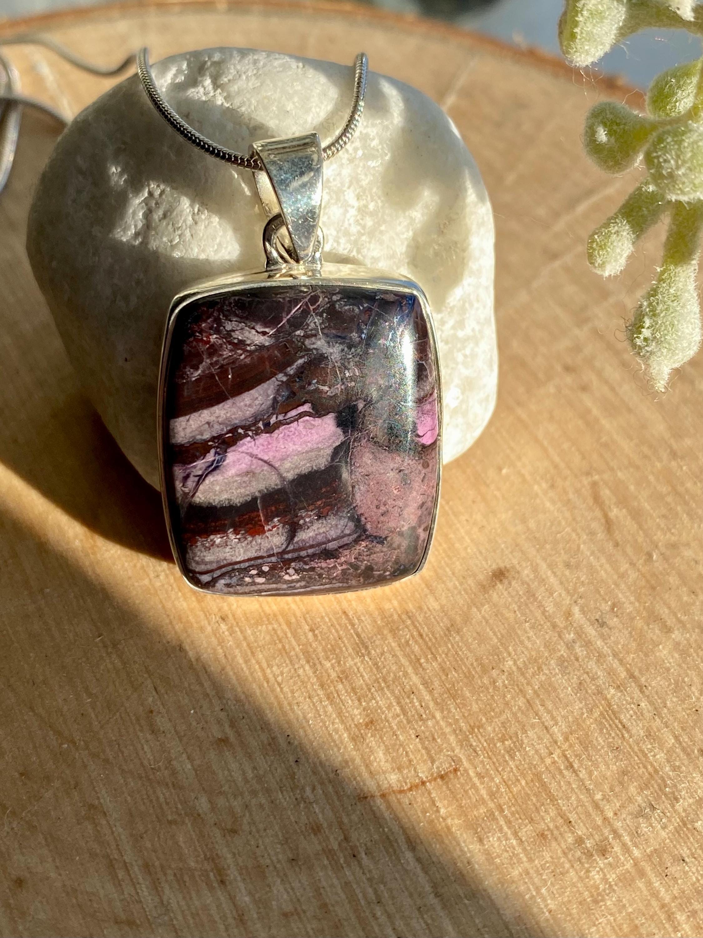 Handmade pendant featuring a rectangular, polished pink, purple and black Sugilite gemstone with natural patterns, bezel-set in 925 sterling silver. The pendant hangs from a delicate silver chain and is photographed in natural sunlight on a wooden surface.