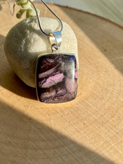 Handmade pendant featuring a rectangular, polished pink, purple and black Sugilite gemstone with natural patterns, bezel-set in 925 sterling silver. The pendant hangs from a delicate silver chain and is photographed in natural sunlight on a wooden surface.