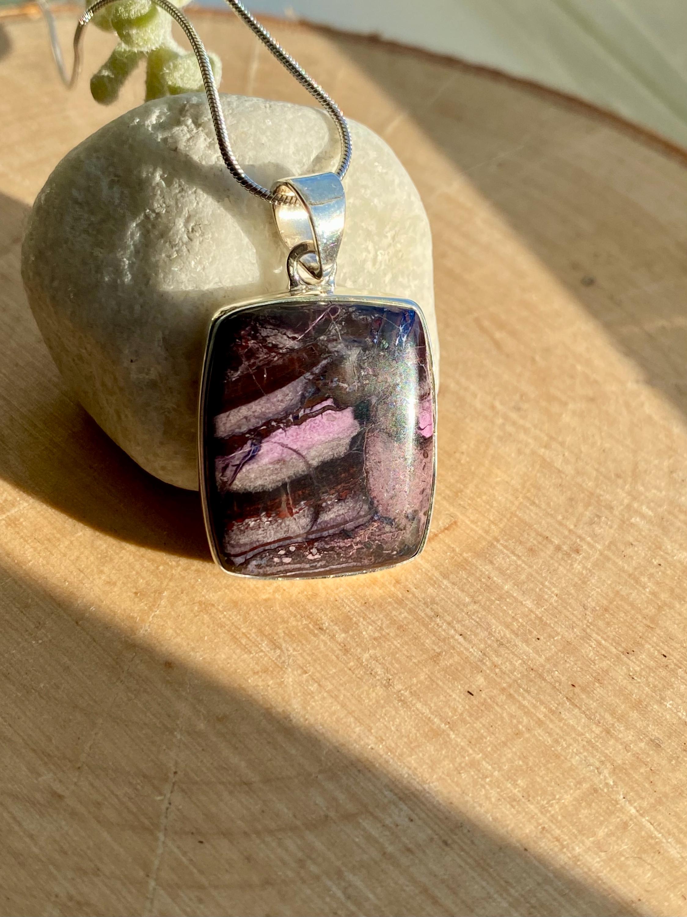 Handmade pendant featuring a rectangular, polished pink, purple and black Sugilite gemstone with natural patterns, bezel-set in 925 sterling silver. The pendant hangs from a delicate silver chain and is photographed in natural sunlight on a wooden surface.
