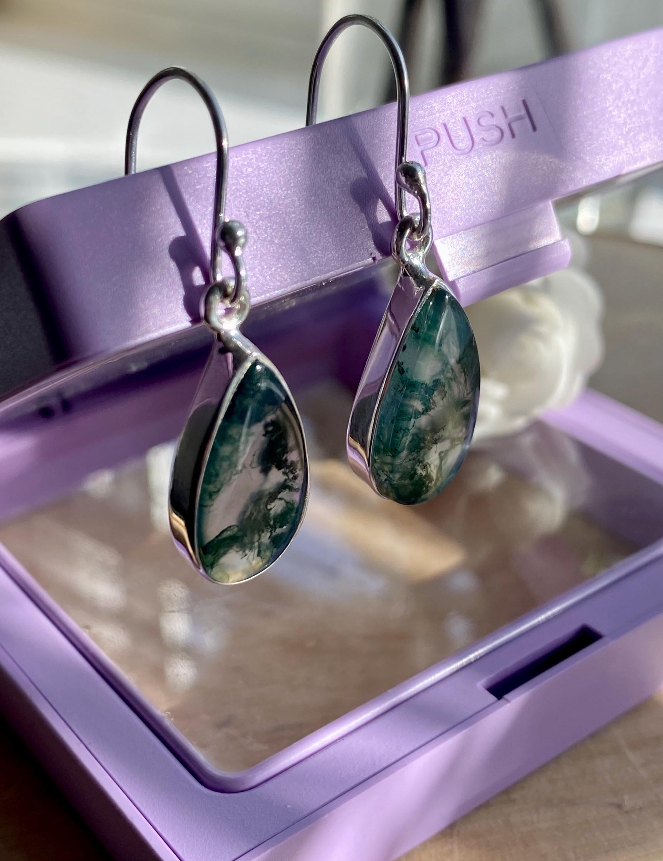 Elegant sterling silver earrings with teardrop-shaped Moss Agate gemstones, featuring natural green inclusions. The bezel-set stones dangle from sleek silver hooks. Displayed on a purple jewellery box in soft natural lighting.