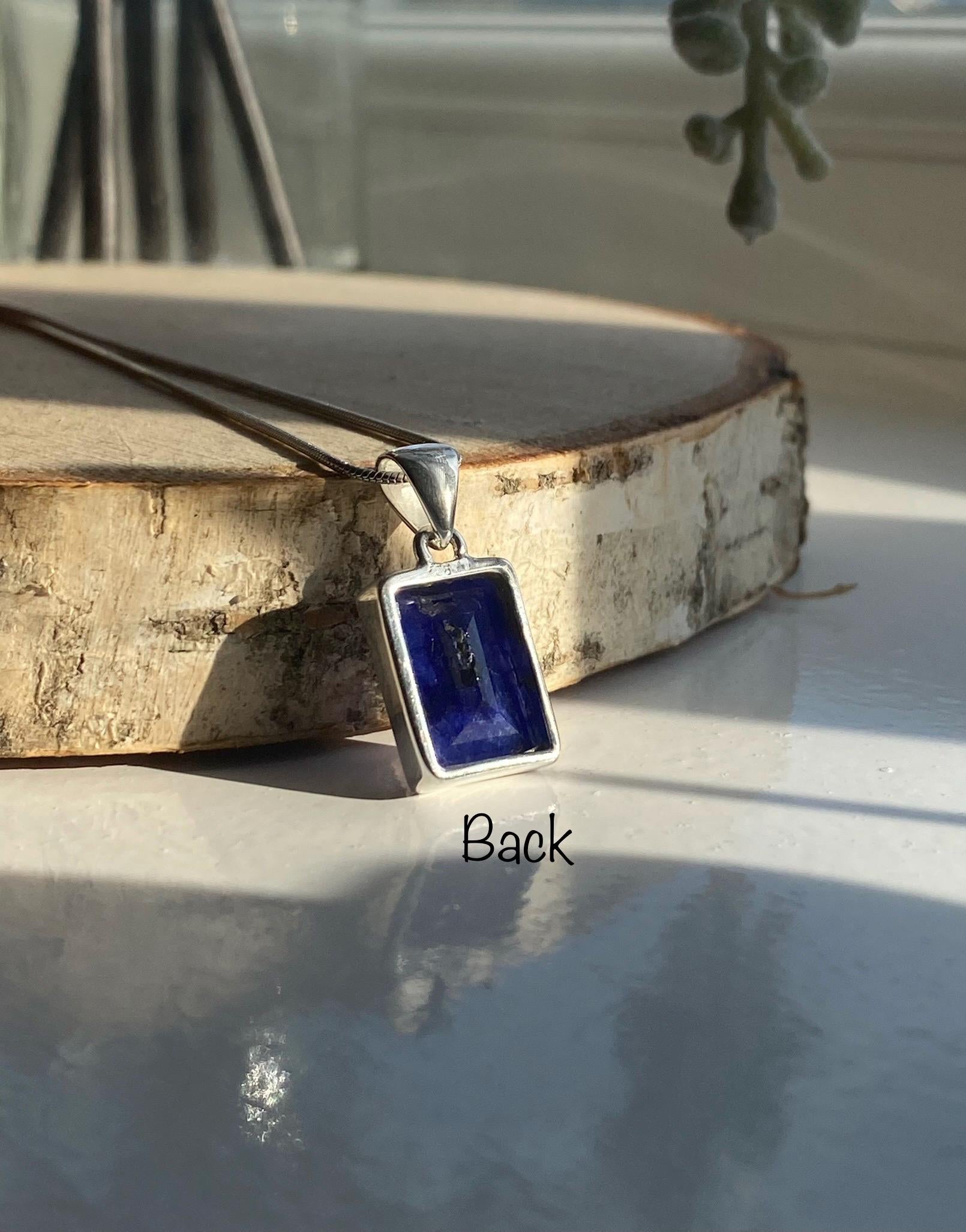Sterling silver pendant featuring a rectangular deep blue Sapphire gemstone with a smooth, polished finish. The pendant is displayed on a wooden slice, with natural sunlight highlighting its rich colour. &#39;Back&#39; text label added for clarity.